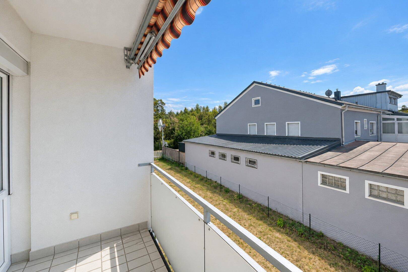 Heller Balkon mit Fliesenboden und Blick auf ein Nachbargebäude und Grünflächen.