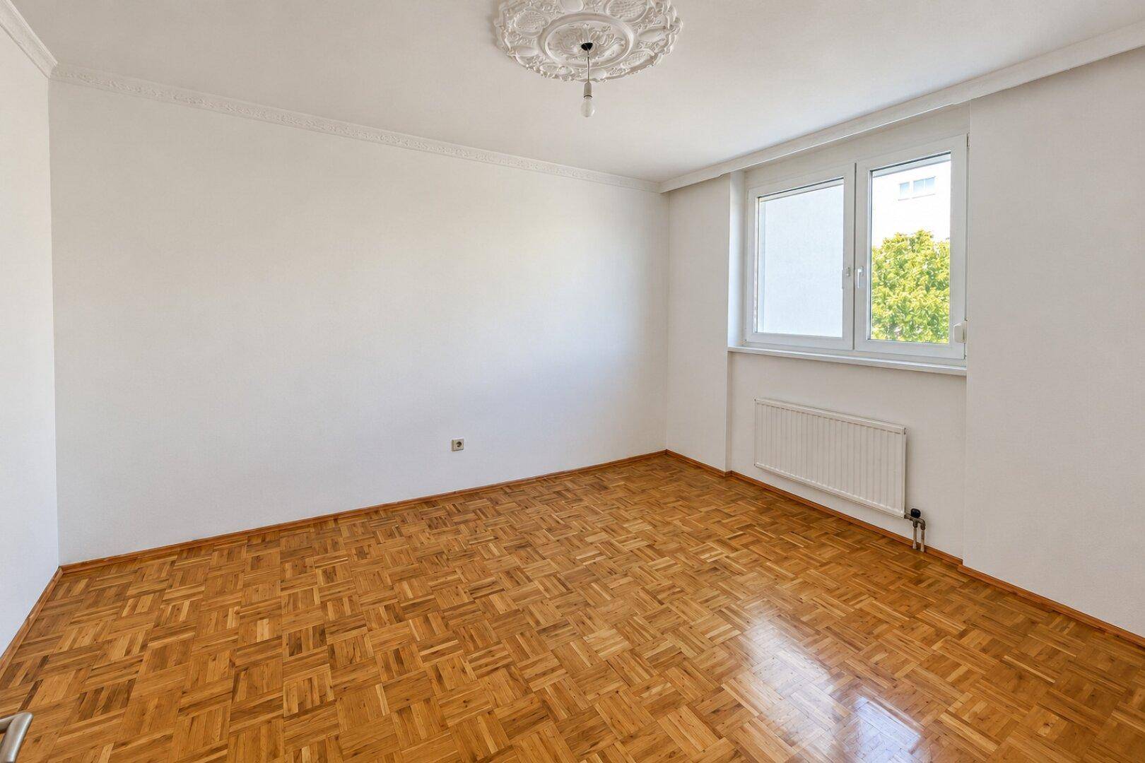 Geräumiges Zimmer mit Parkettboden, weißen Wänden und dekorativer Deckenrosette.