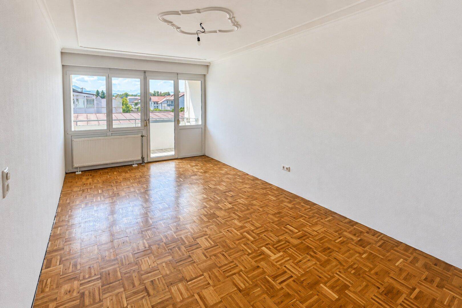 Geräumiges Wohnzimmer mit Parkettboden, Balkonzugang und dekorativer Deckenrosette.