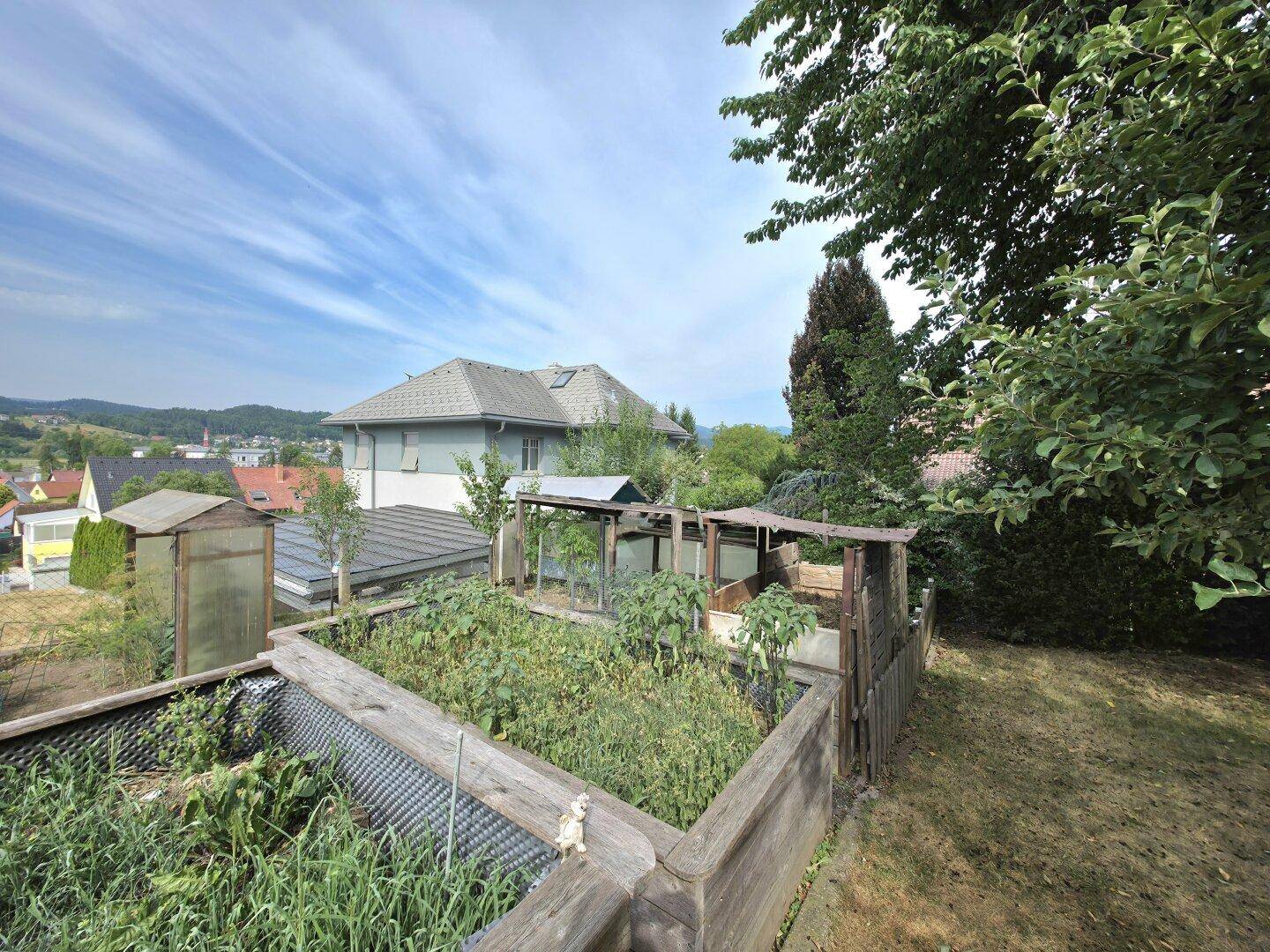 Gartenbereich mit Hochbeeten und kleinen Gewächshäusern, umgeben von Bäumen und Sträuchern.