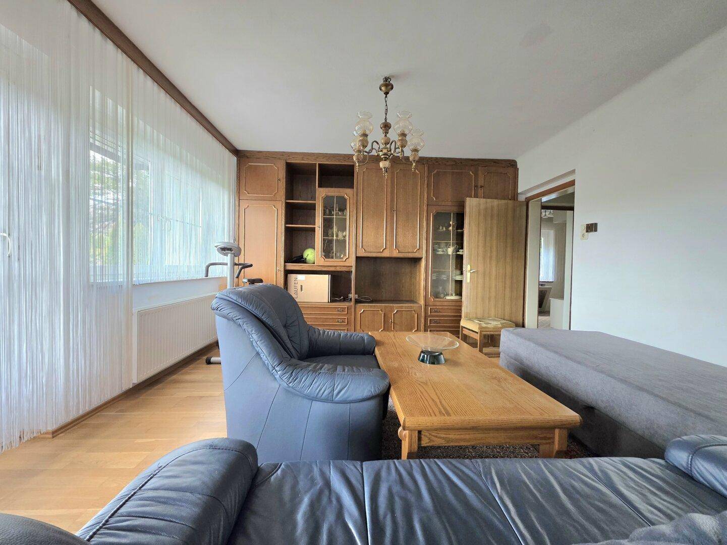 Gemütliches Wohnzimmer mit Holzmöbeln, bequemen Sofas und großen Fenstern mit Vorhängen.