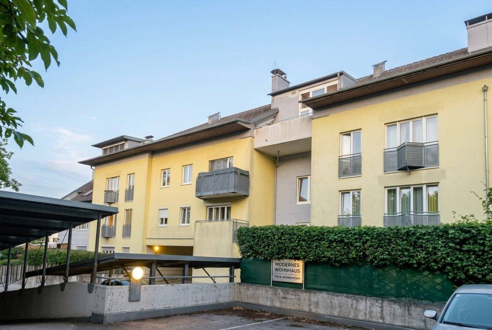 Mehrfamilienhaus mit gelber Fassade, Balkonen und überdachten Parkplätzen.