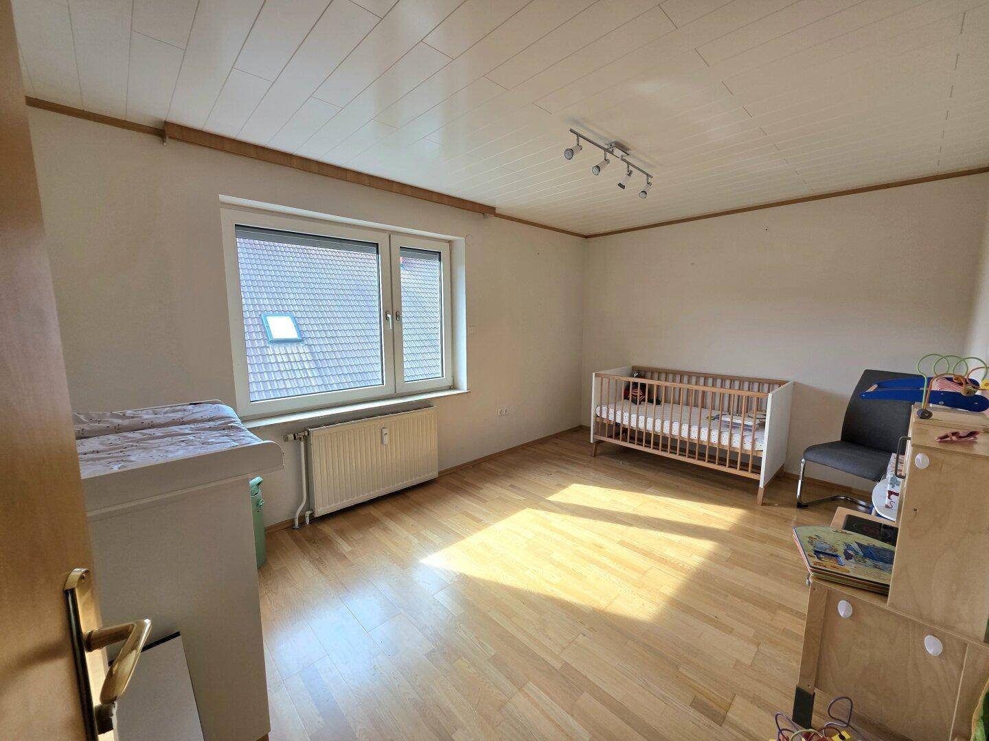 Helles Kinderzimmer mit Holzboden, Babybett und Fenster mit Blick auf das Dach.