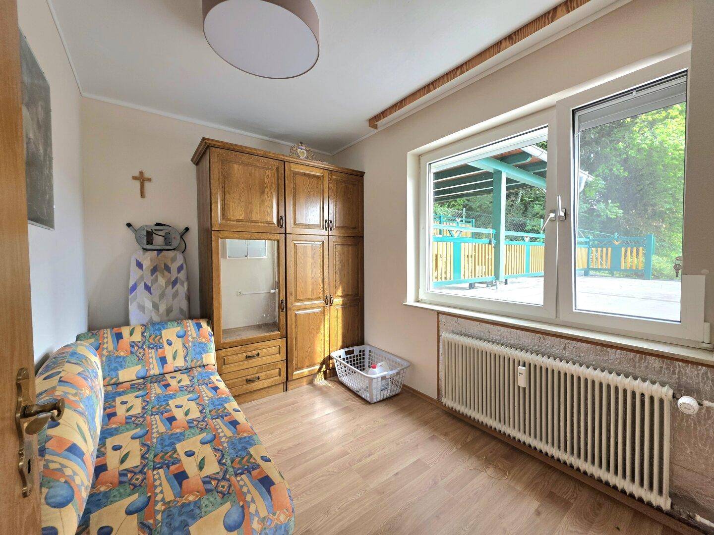Schlafzimmer mit Holzboden, Holzschrank und Fenster mit Blick auf den überdachten Außenbereich.