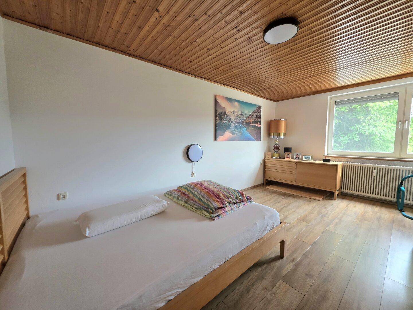 Helles Schlafzimmer mit Holzboden, Holzbett und Fenster mit Blick ins Grüne.