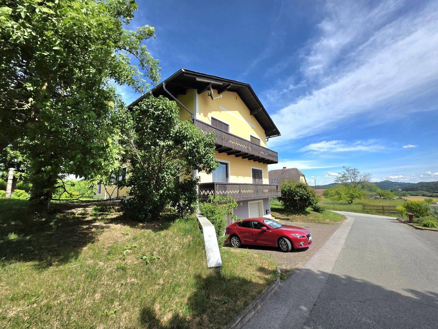 Gelbes Haus mit Balkonen und einem roten Auto auf der Zufahrt, umgeben von grüner Landschaft.
