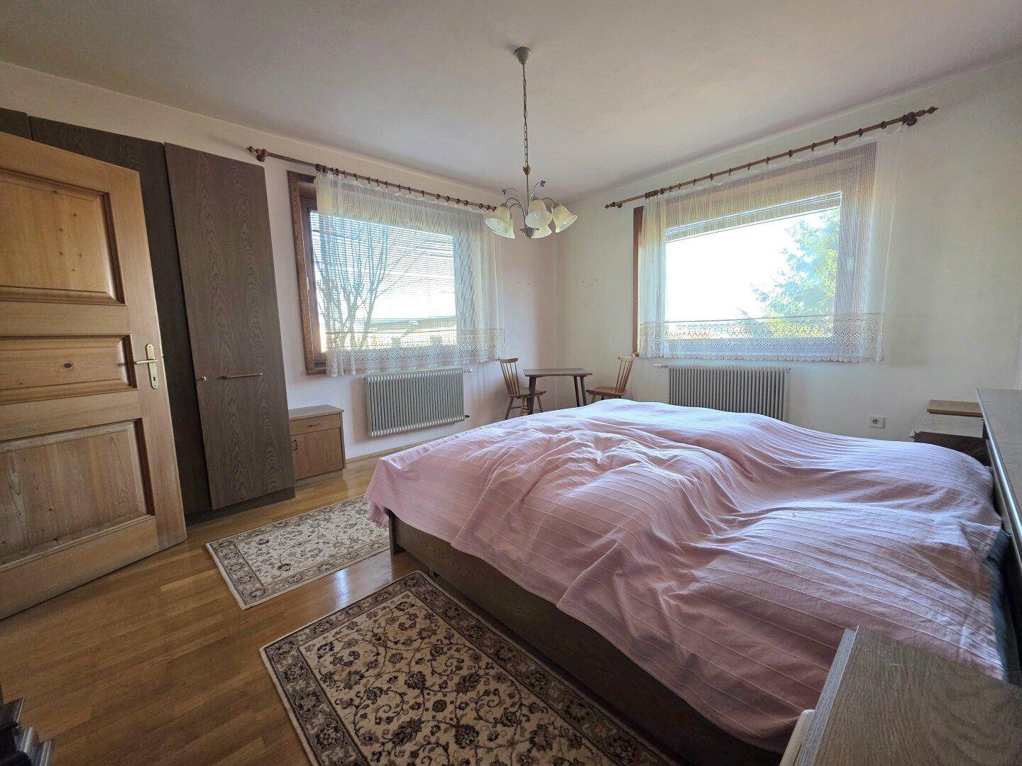 Gemütliches Schlafzimmer mit Doppelbett, Holzschrank und zwei Fenstern, die viel Licht hereinlassen.