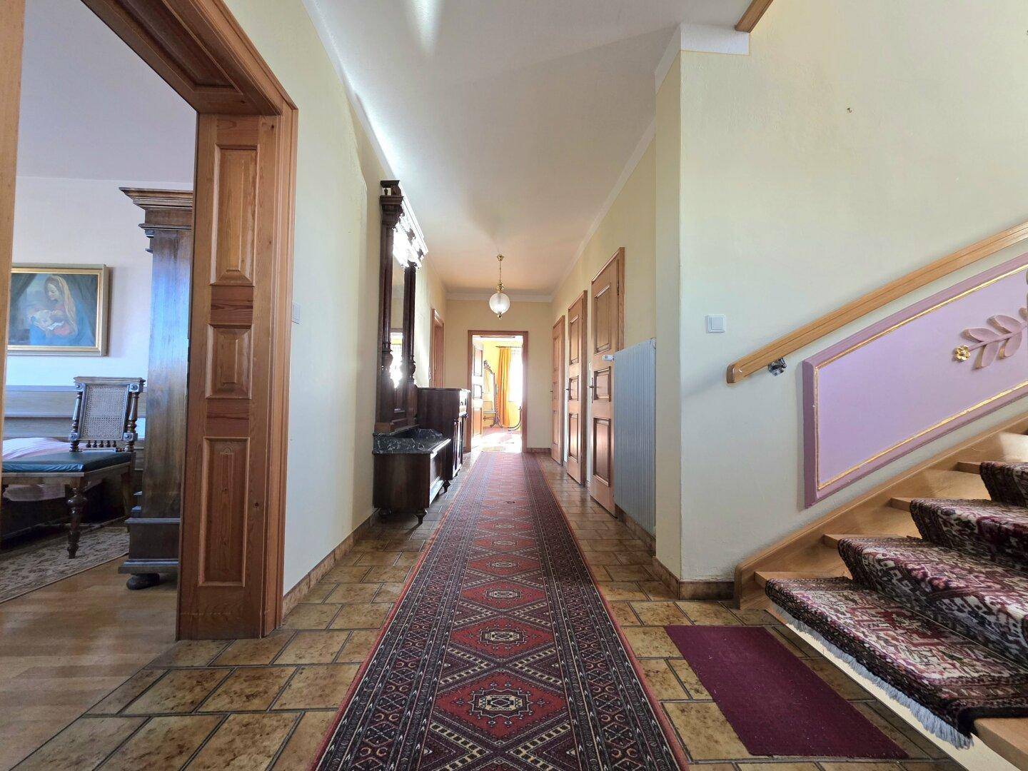 Langer Flur mit gemustertem Teppich, Holzmöbeln und einer Treppe mit dekorativen Elementen.
