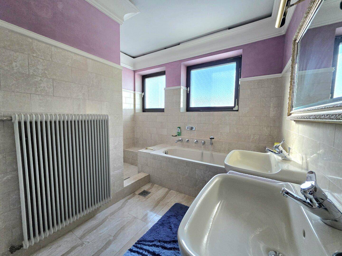 Badezimmer mit zwei Waschbecken, Badewanne und hellen Fliesen, kombiniert mit lila Wänden.