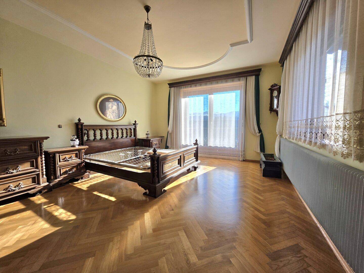 Elegantes Schlafzimmer mit dunklem Holzparkett, Kronleuchter und großen Fenstern, die den Raum erhellen.