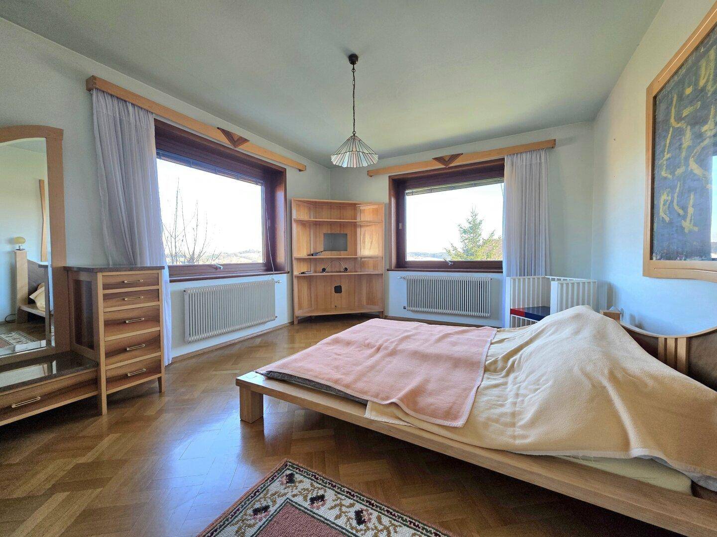 Helles Schlafzimmer mit Holzbett, Kommode und zwei großen Fenstern, die viel Tageslicht bieten.