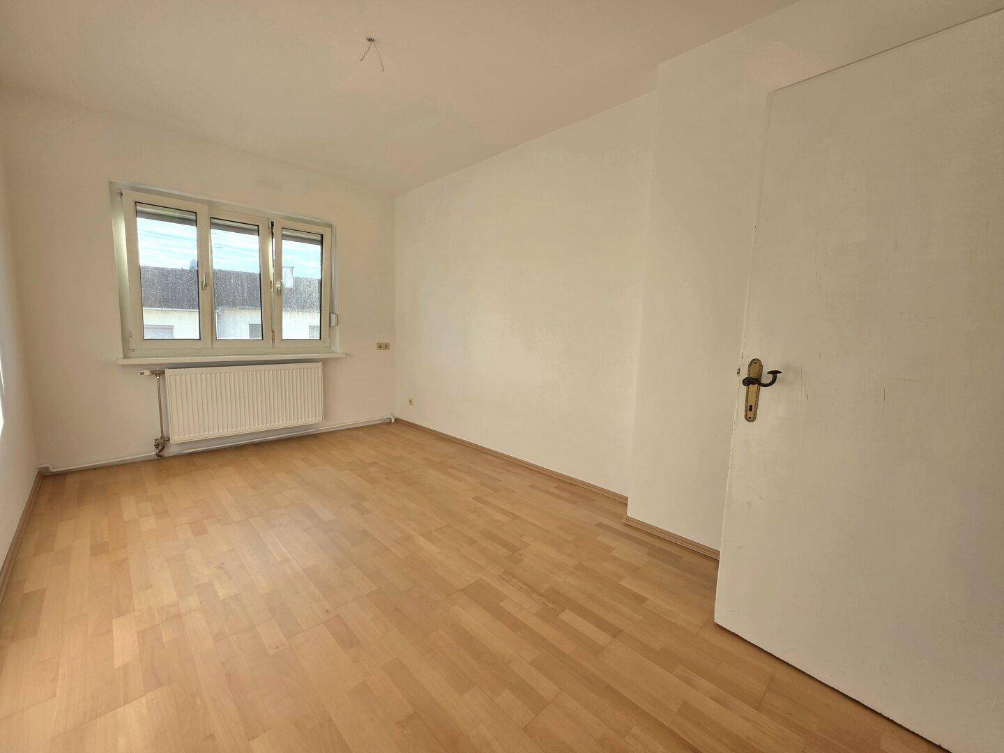 Helles Schlafzimmer mit Parkettboden, weißen Wänden und einem Fenster, das Tageslicht spendet.