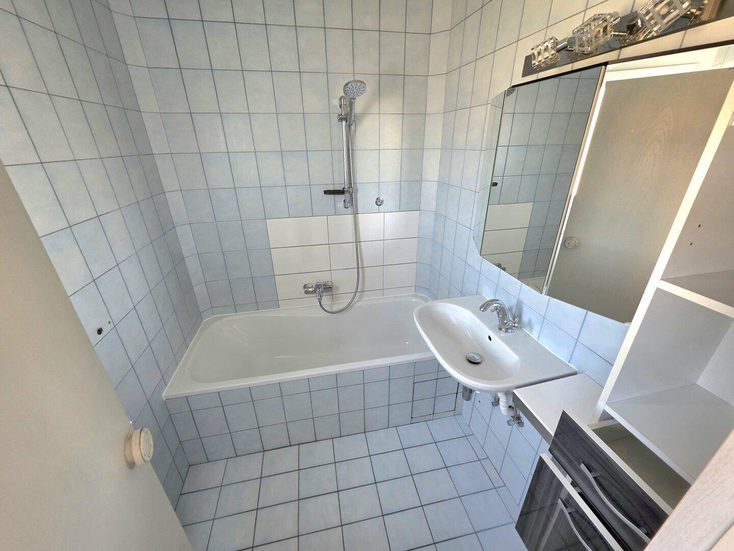 Badezimmer mit hellblauen Fliesen, Badewanne mit Duschfunktion und einem Waschbecken mit Spiegelschrank.
