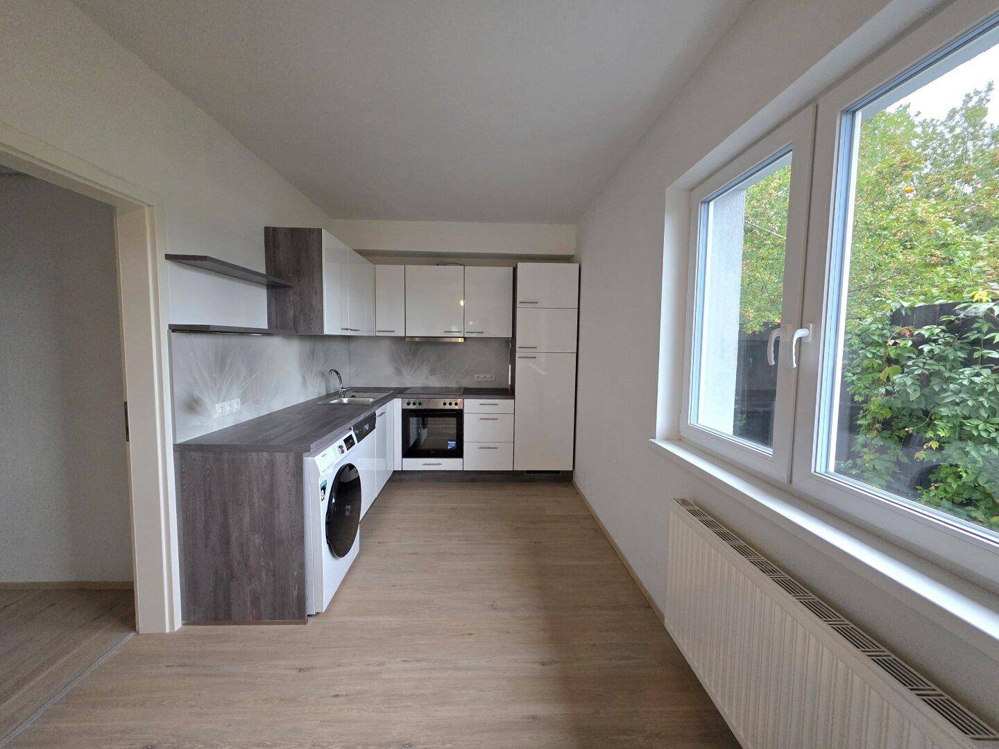 Helle, moderne Küche mit weißen und grauen Schränken, integrierter Waschmaschine und großem Fenster.