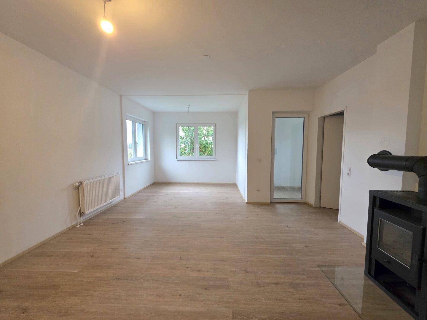 Geräumiges, leeres Wohnzimmer mit hellem Laminatboden, großen Fenstern und einem modernen Kaminofen.