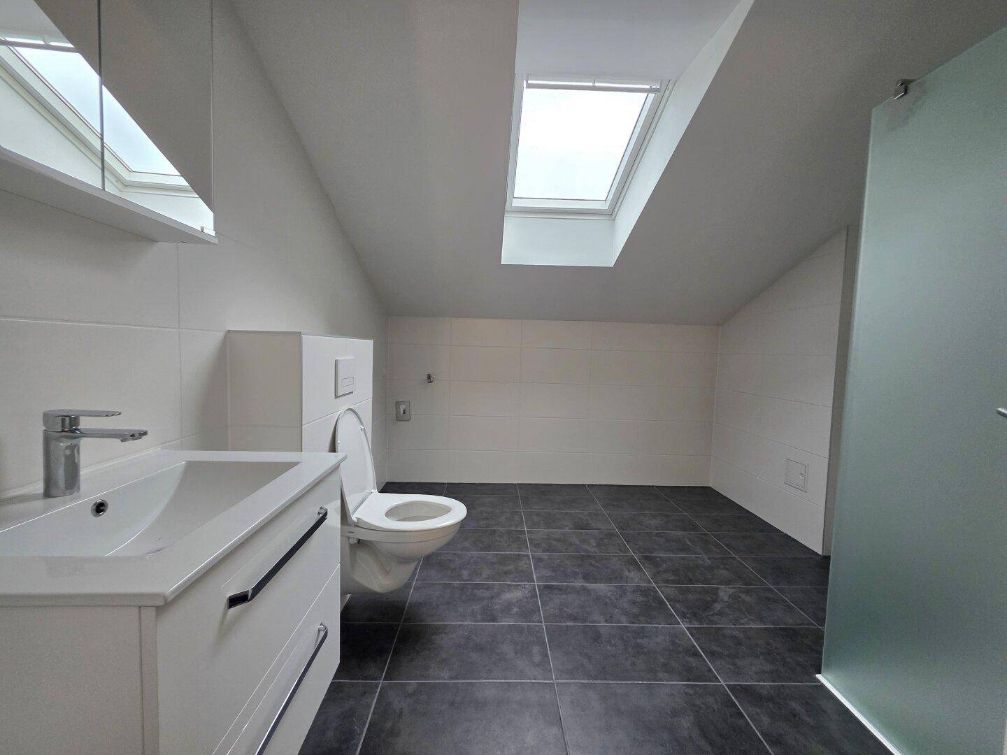 Modernes Badezimmer im Dachgeschoss mit dunklen Fliesen, wandhängendem WC, Waschtisch und Dachfenster.