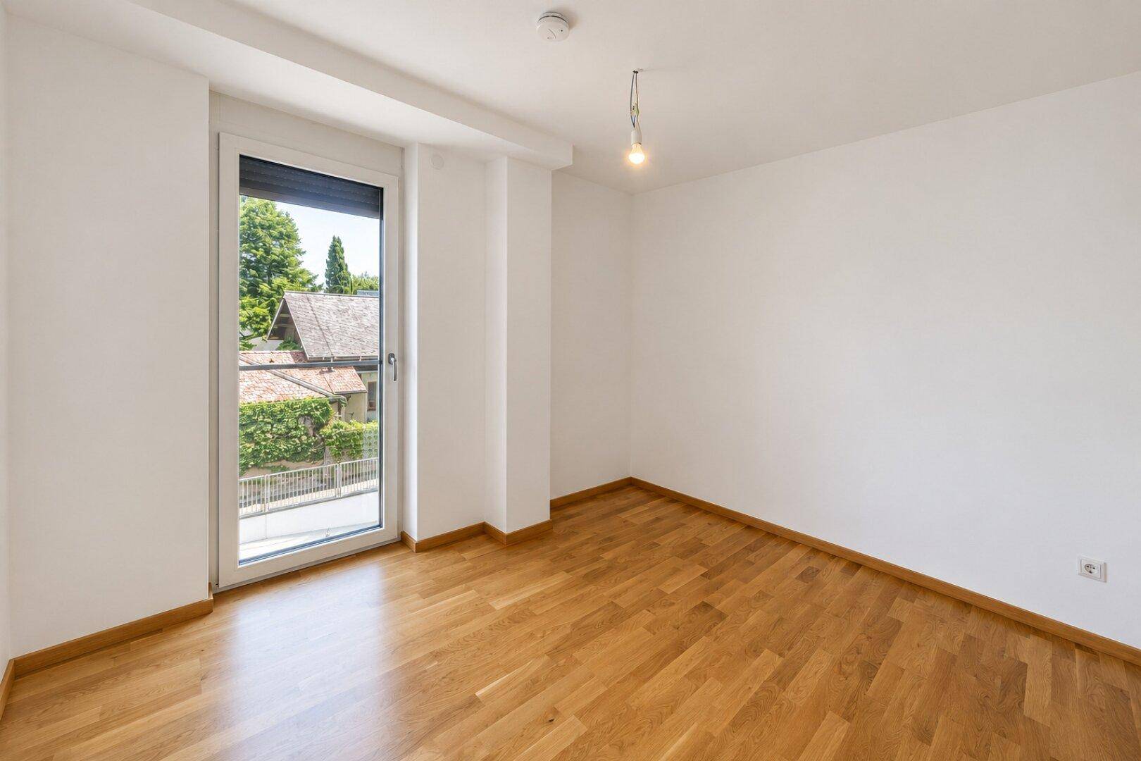 Schlafzimmer mit Parkettboden und bodentiefem Fenster, das viel Licht hereinlässt.