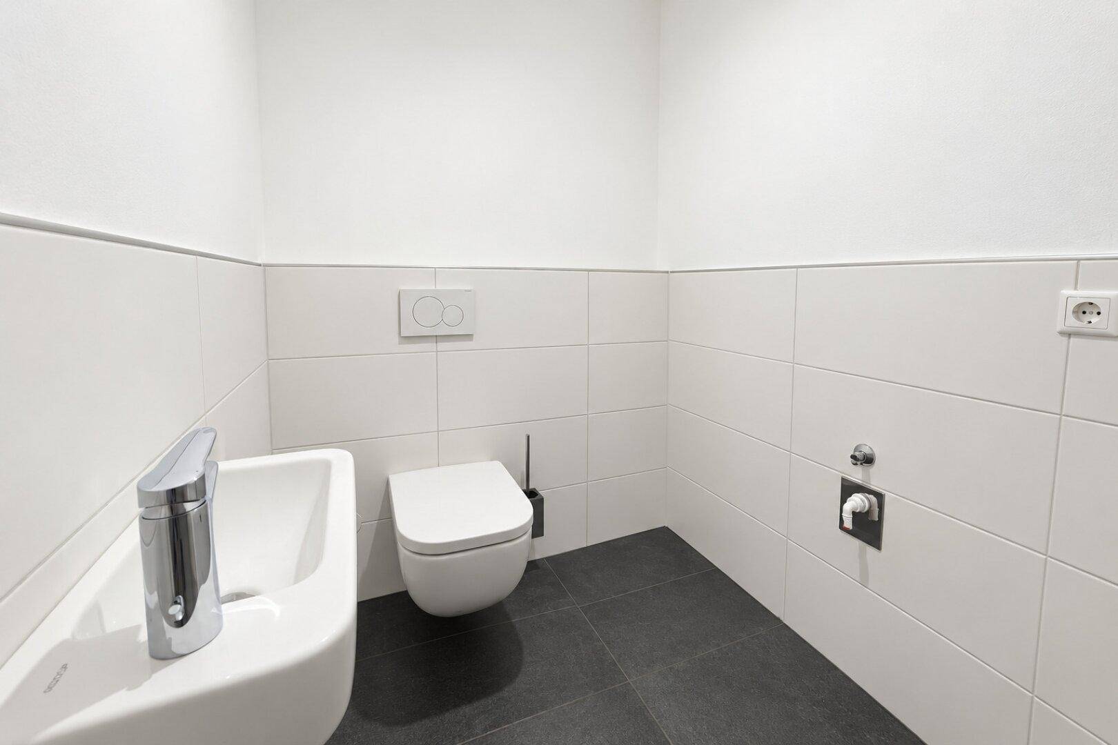 Modernes Gäste-WC mit weißen Wandfliesen, dunklem Boden und wandhängendem WC sowie kleinem Waschbecken.