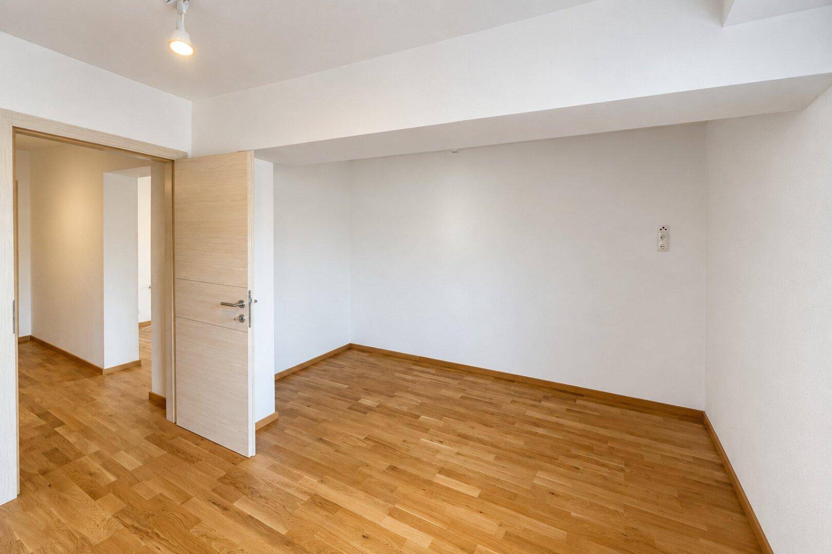 Helles, leeres Zimmer mit Parkettboden und weiß gestrichenen Wänden, ideal für Schlaf- oder Arbeitszimmer.