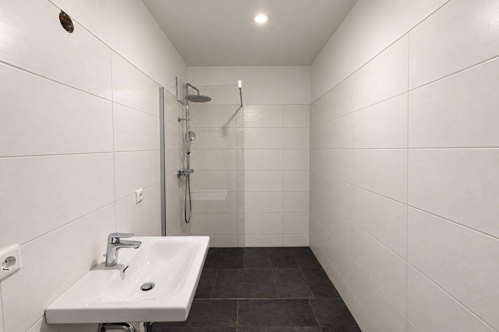 Helles Badezimmer mit modernem Waschbecken, ebenerdiger Dusche und weißen Fliesen an Wänden und Boden.