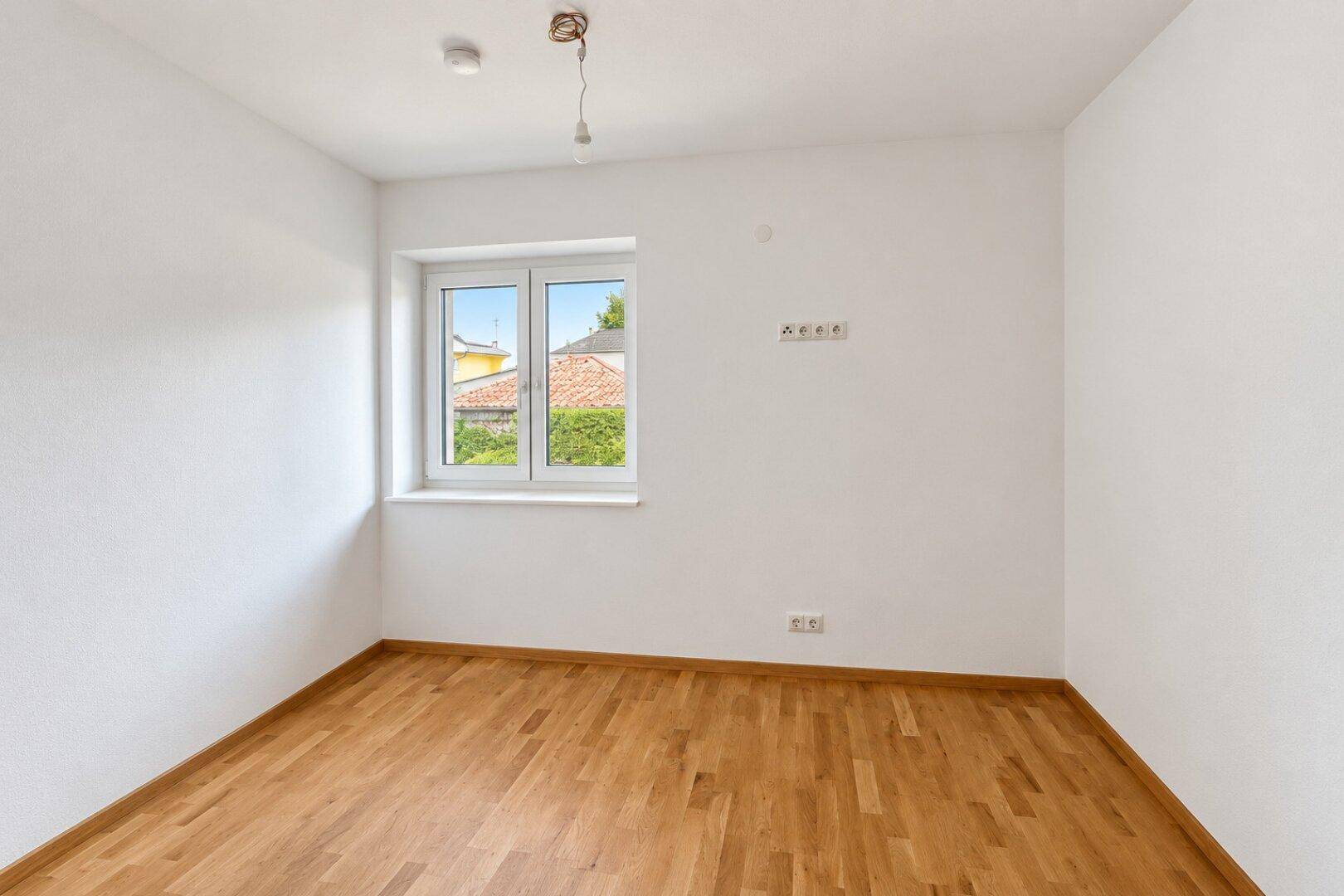 Helles Zimmer mit Parkettboden und Fenster, das einen grünen Ausblick auf die Umgebung bietet.