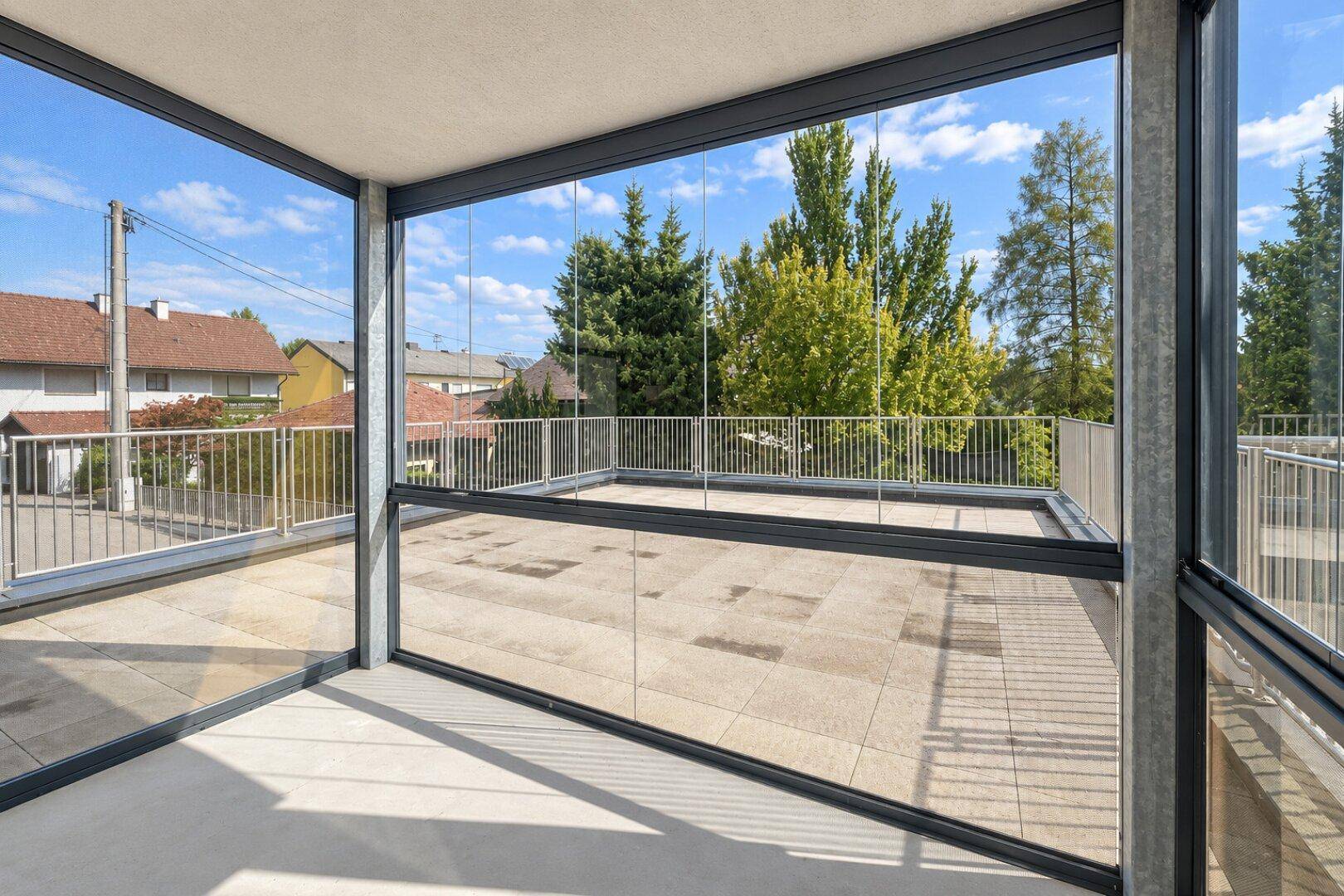 Verglaste Loggia mit weitem Blick auf die Umgebung und die angrenzende Terrasse, umgeben von Bäumen.