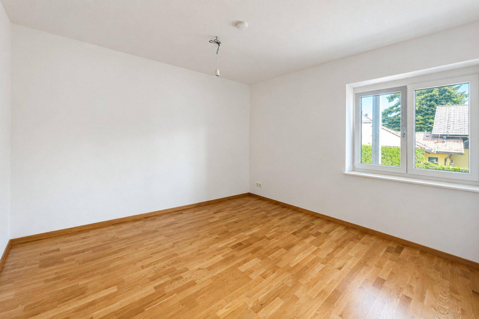 Freundliches Zimmer mit hellem Parkettboden und Fenster mit Blick ins Grüne, ideal für Ruhe und Entspannung.