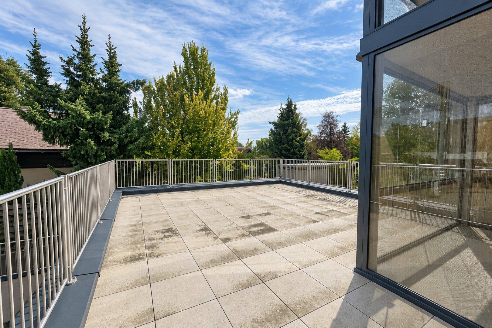 Weitläufige Dachterrasse mit Fliesenboden und Panoramablick auf die grüne Umgebung und umliegende Häuser.