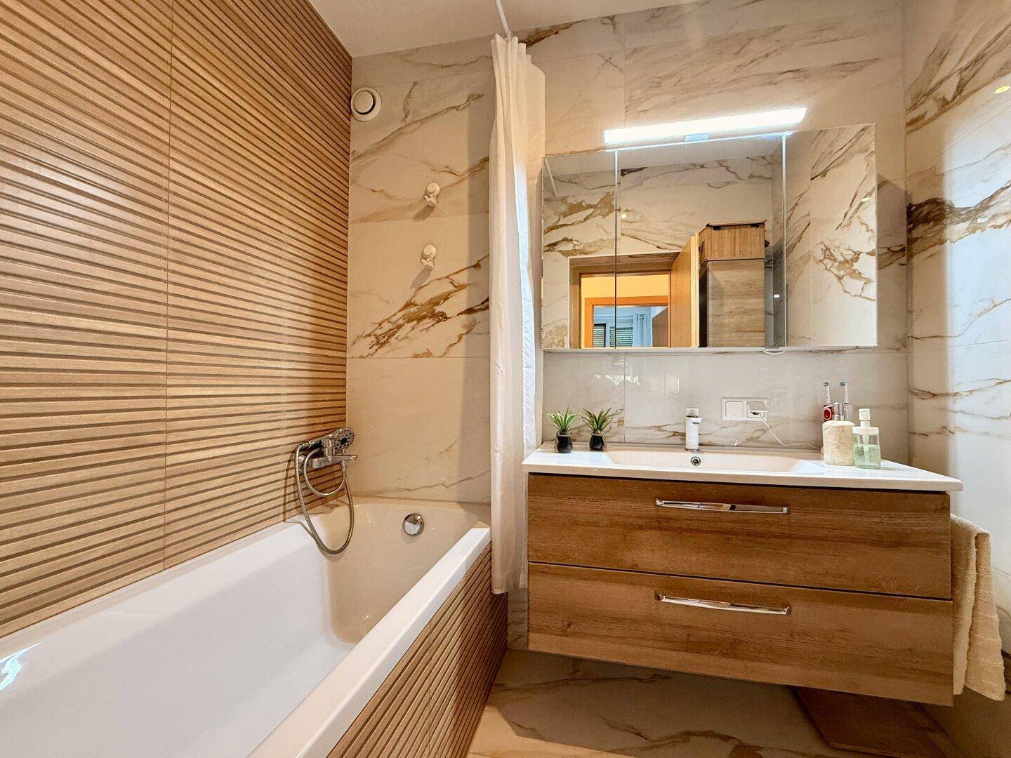Modernes Badezimmer mit Badewanne, Holzlamellenwand und Marmor-Effektfliesen.