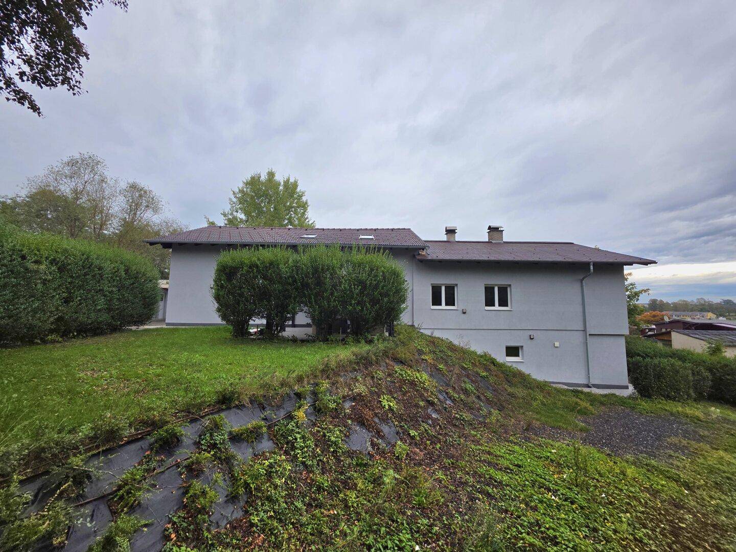 Seitenansicht des grauen Hauses mit gepflegtem Garten, Hecken und bewachsenem Hang.