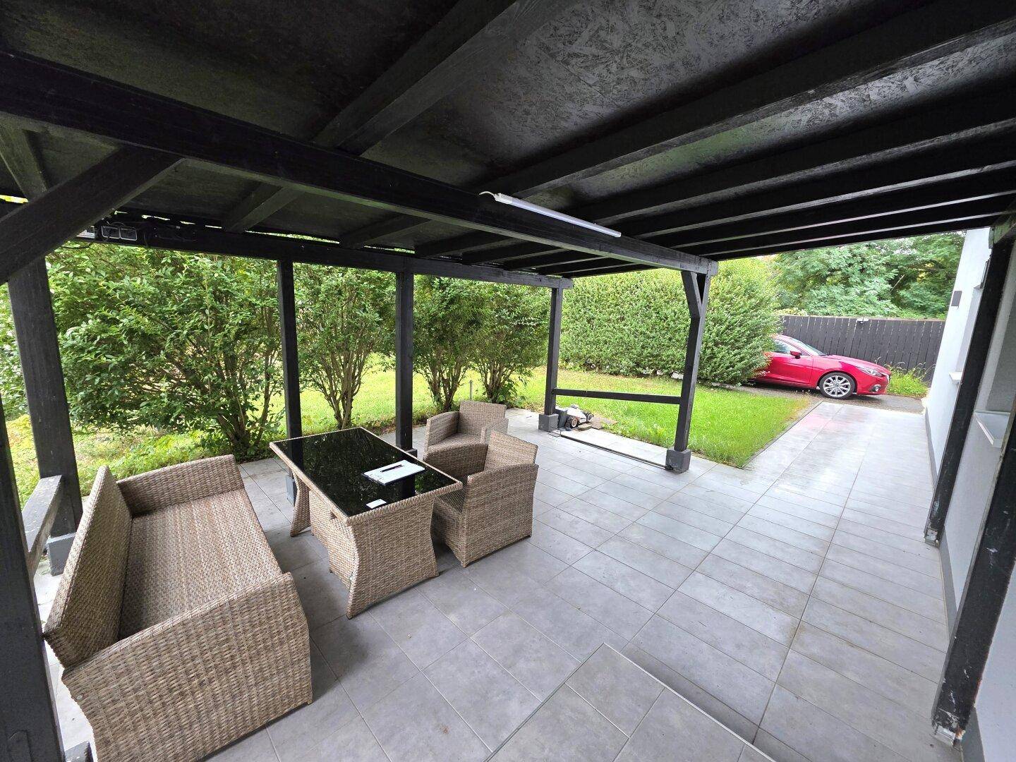 Überdachte Terrasse mit Rattanmöbeln und Blick auf den Garten sowie die Einfahrt mit Auto.