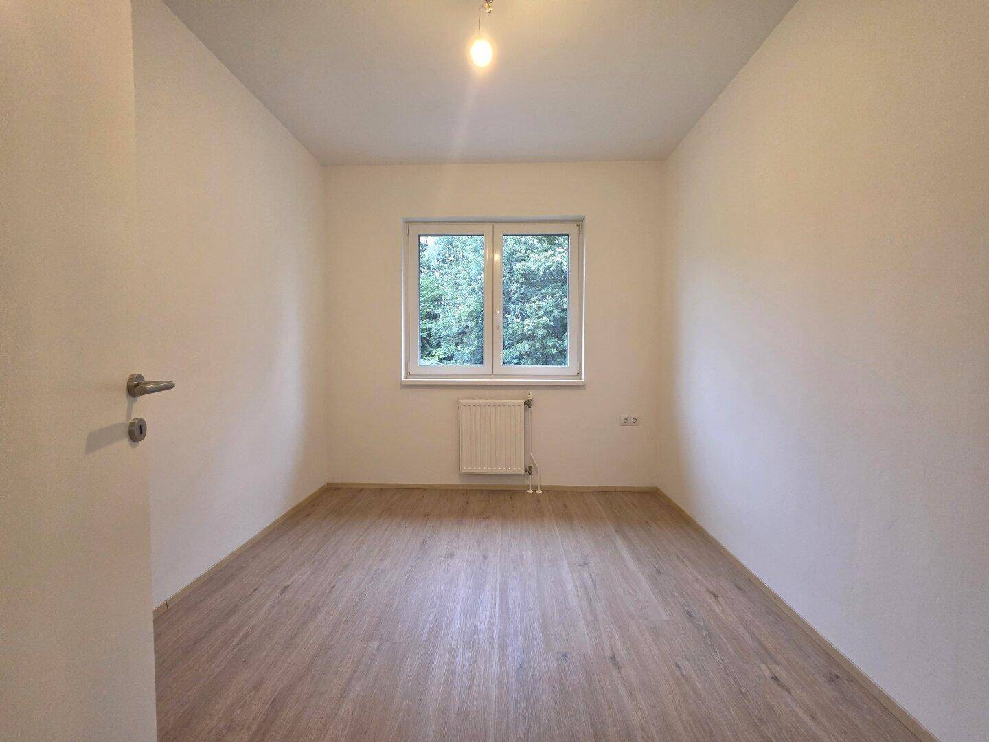 Helles Schlafzimmer mit Laminatboden, weiß gestrichenen Wänden und Fenster mit Blick ins Grüne.