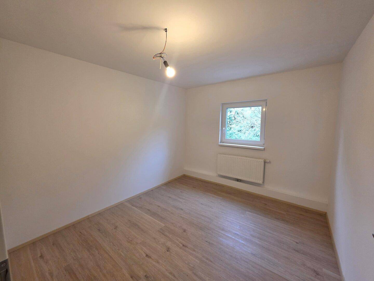 Helles Schlafzimmer mit Laminatboden, weiß gestrichenen Wänden und Fenster mit Blick ins Grüne.