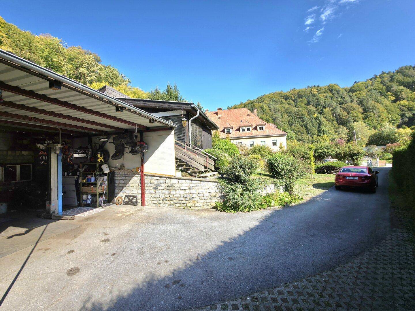 Außenansicht mit Garage, Werkstattbereich und Auffahrt, umgeben von grünen Hügeln und Bäumen.