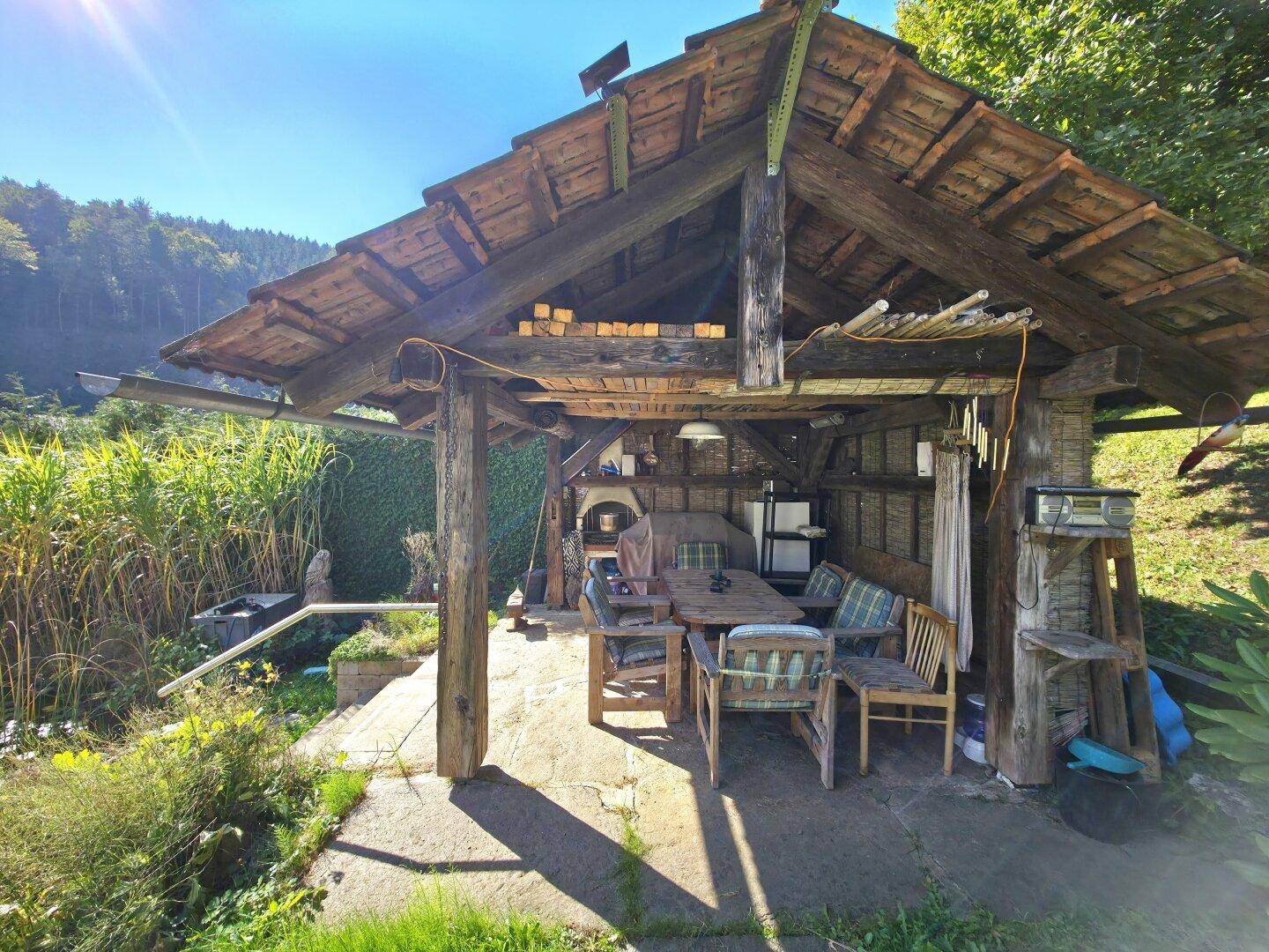 Rustikaler Holzpavillon mit Esstisch und Stühlen, ideal für Mahlzeiten im Freien, umgeben von Natur.