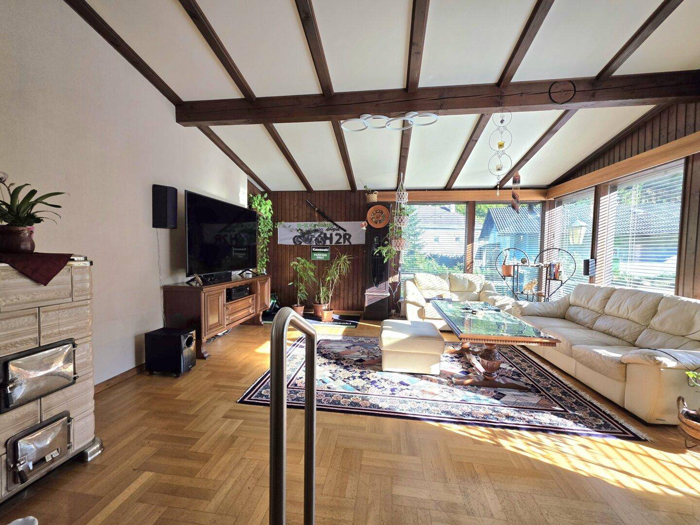Helles Wohnzimmer mit Holzbalkendecke, Parkettboden und großen Fenstern, die viel Licht hereinlassen.