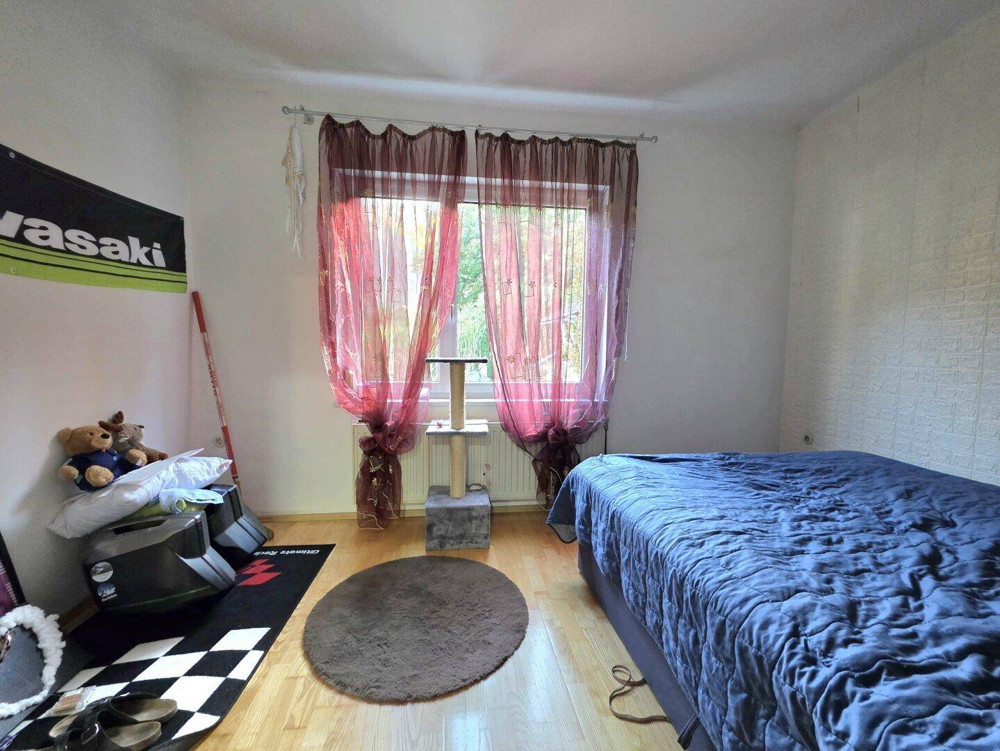 Gemütliches Schlafzimmer mit blauem Bett, Holzfußboden und einem Fenster mit roten Vorhängen.