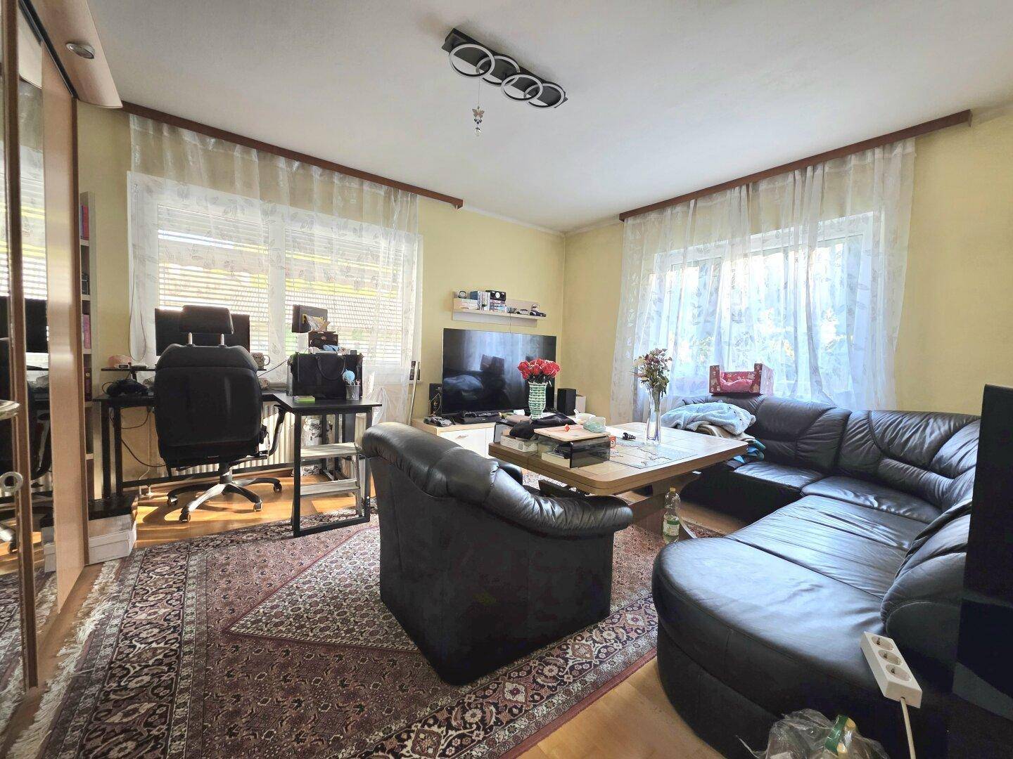 Wohnzimmer mit großem Ecksofa, Schreibtisch und Teppich, hell durch die Fenster mit weißen Vorhängen.