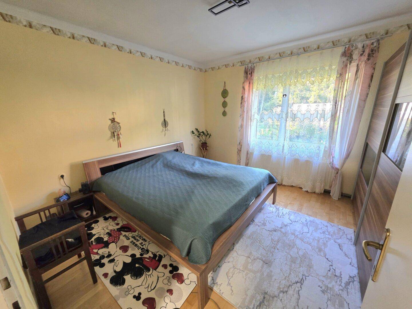 Schlafzimmer mit Holzrahmenbett, hellgelben Wänden und einem Fenster mit floralen Vorhängen.