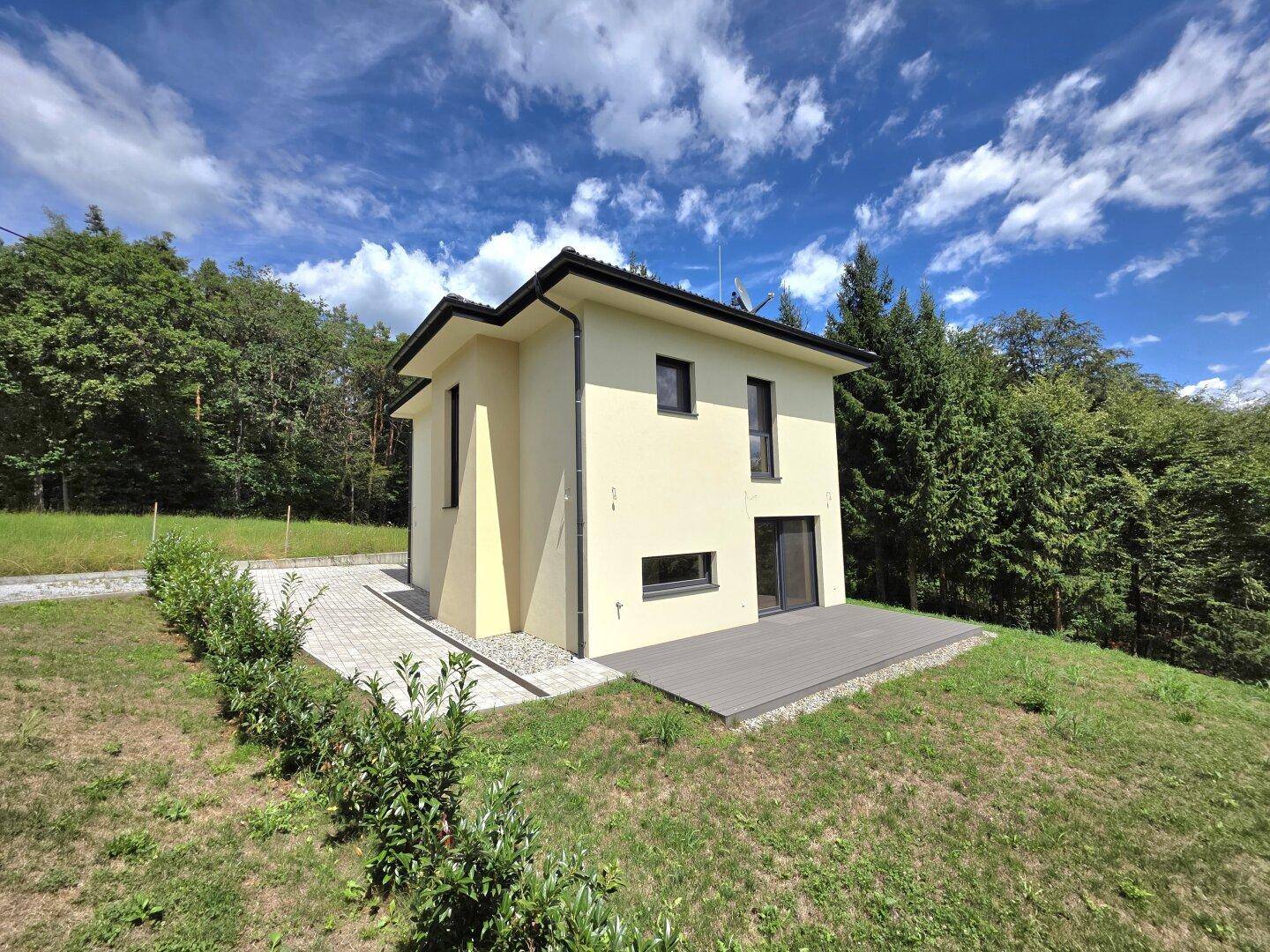 Modernes Einfamilienhaus mit heller Fassade und Holzterrasse, umgeben von viel Grün und blauem Himmel.