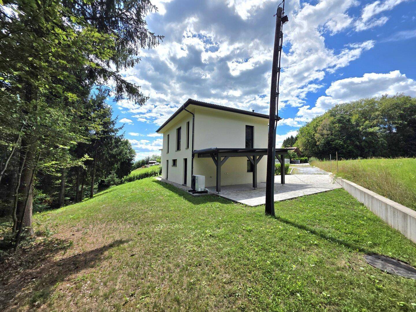 Rückansicht eines modernen Hauses mit überdachtem Bereich und weitläufiger Grünfläche unter blauem Himmel.