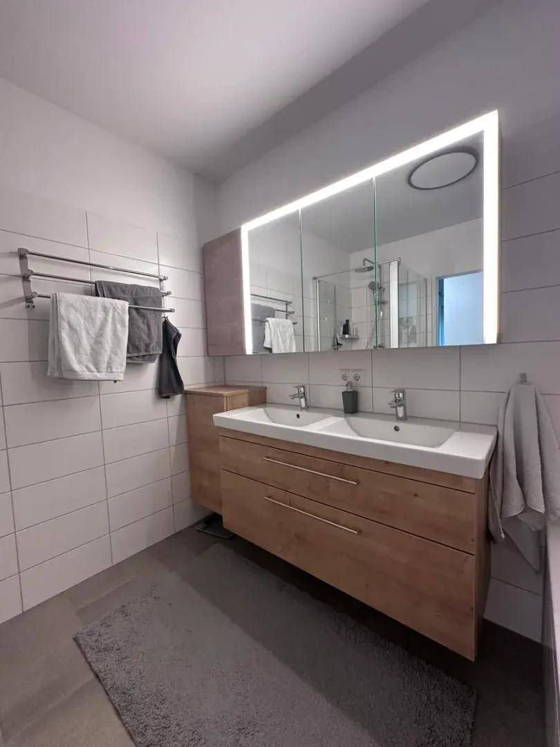 Modernes Badezimmer mit Doppelwaschbecken, Holzunterschrank und beleuchtetem Spiegelschrank.