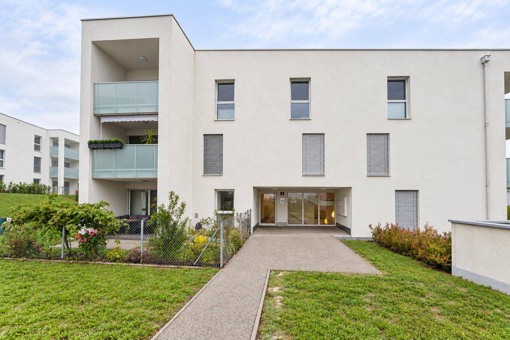 Moderne Fassade des Apartmentgebäudes mit Eingangsbereich, Balkonen und gepflegter Grünfläche.
