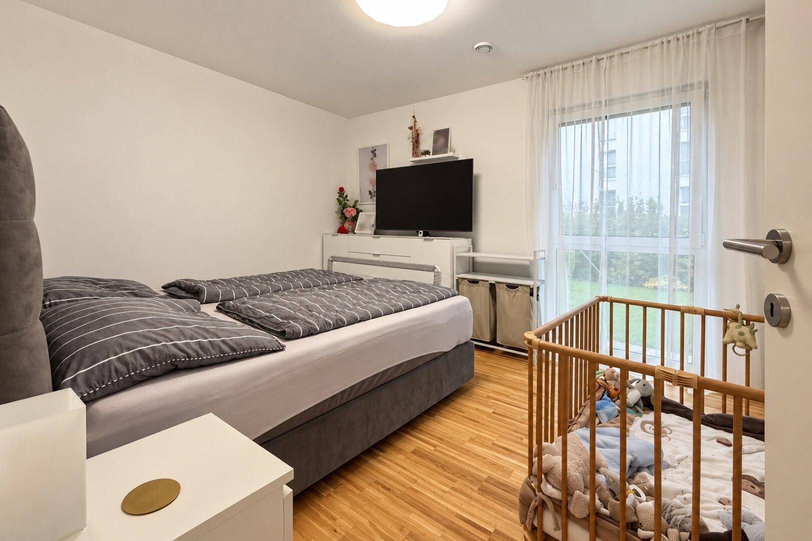 Helles Schlafzimmer mit Holzboden, Doppelbett, Babybett und großem Fenster mit Blick ins Grüne.