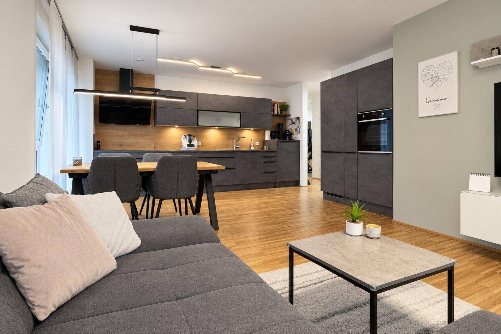 Offener Wohn- und Essbereich mit gemütlichem Sofa und integrierter, moderner Küche im Hintergrund.