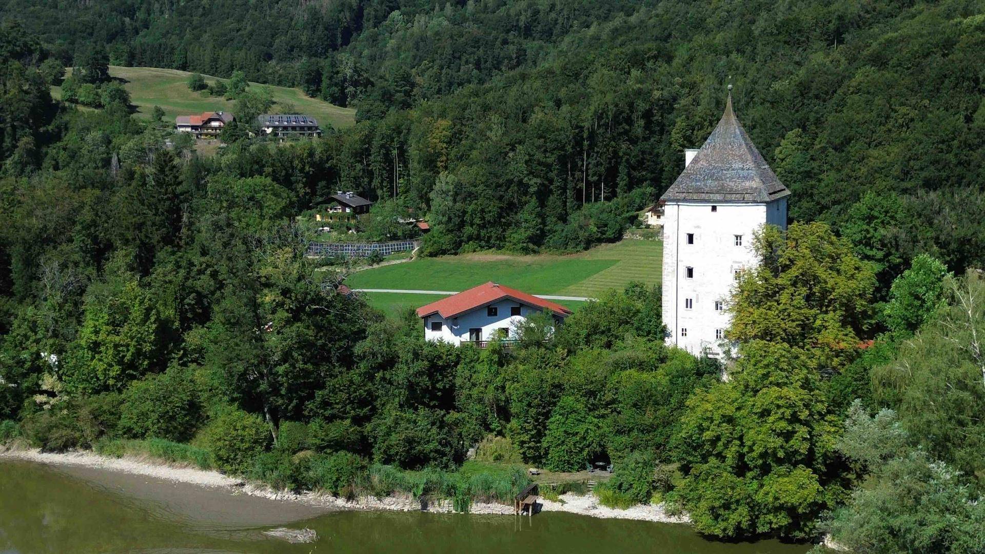 Malerische Landschaft mit historischem Turmgebäude und einem Haus am Flussufer, umgeben von Wäldern.