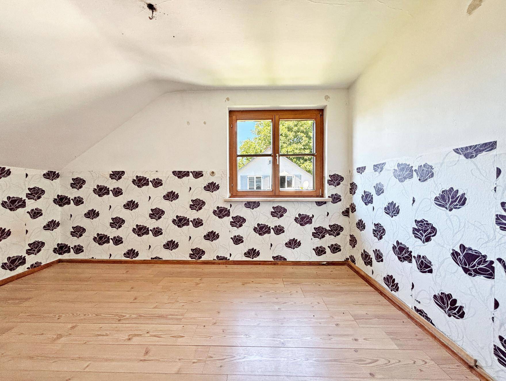 Dachgeschosszimmer mit Holzdielenboden, schrägen Wänden und floraler Tapete an den unteren Wänden.