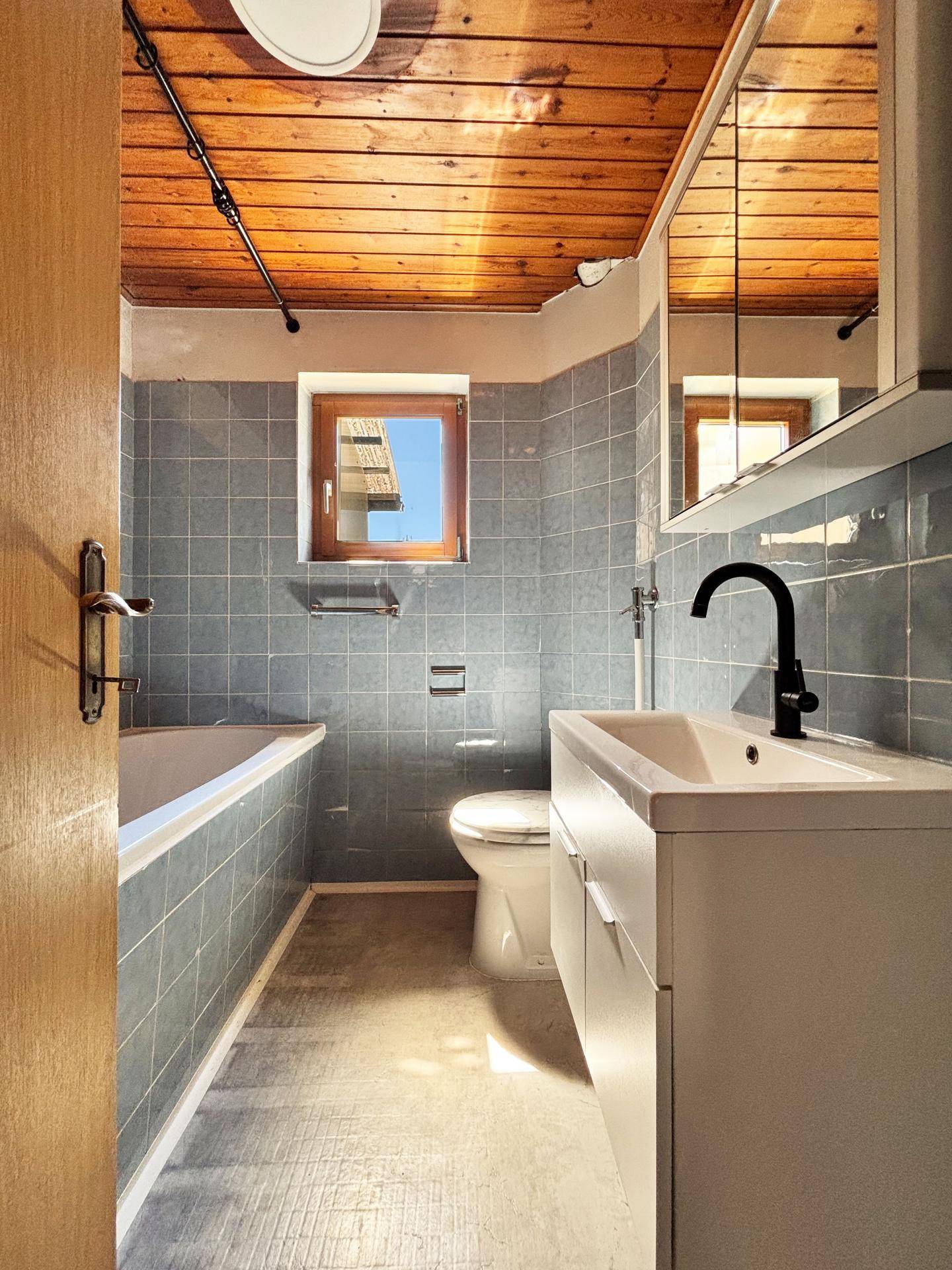 Badezimmer mit Badewanne, Waschbecken und WC, ausgestattet mit blauen Fliesen und Holzdecke.