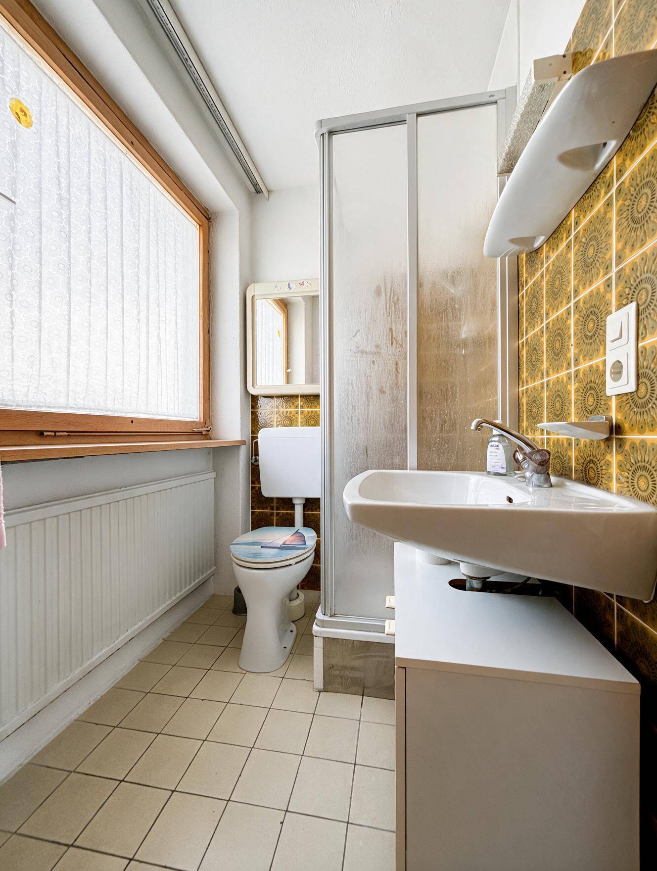 Badezimmer mit Dusche, WC, Waschbecken und Fenster, ausgestattet mit 80er-Jahre-Fliesen und viel Licht.