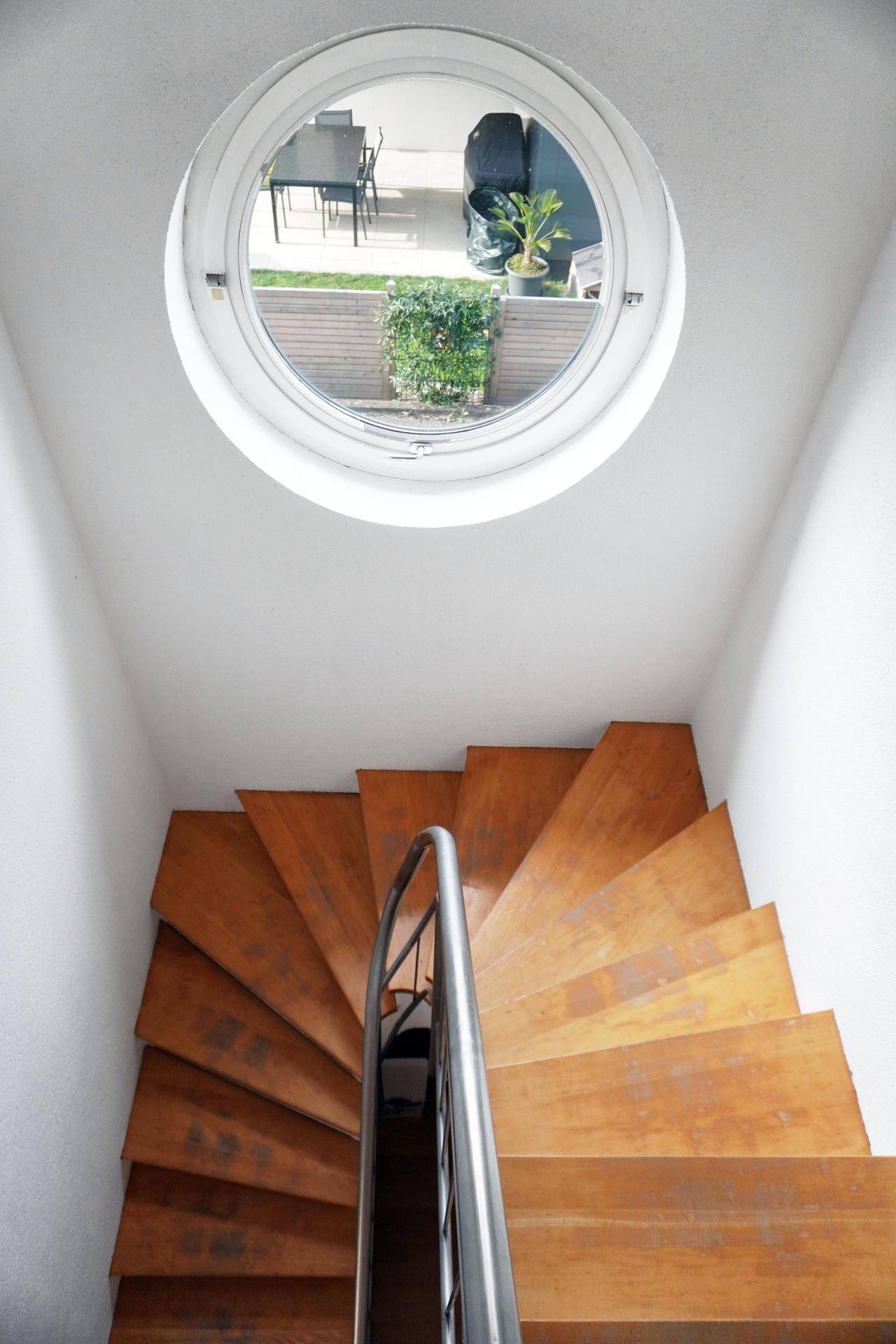 Holztreppe mit Metallgeländer und einem runden Fenster, das Licht hereinlässt.